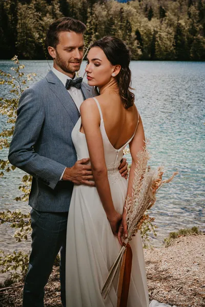 Die Braut mit Blumen auf dem Rücken im Arm des Bräutigams am Ufer des Sees